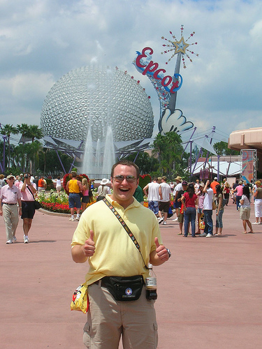man wearing fanny pack and tourist clothes smiles in front of disney epcot center and crowd
